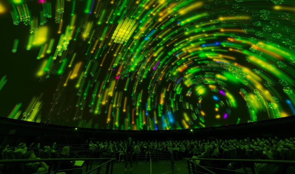 Green star trails track across the dome in a planetarium will a full audience.