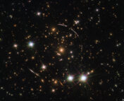A field of galaxies on a black background, with a spherical orange galaxy at the center of the image. Thin short bright arcs trace a circle centered on the orange galaxy.