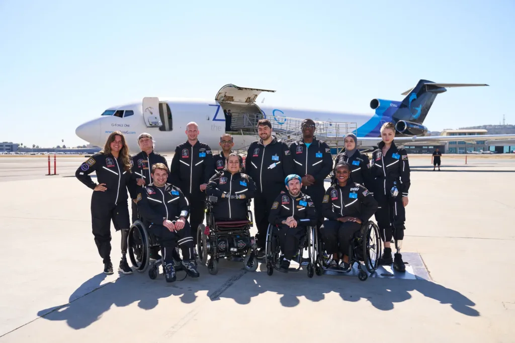 8 people standing with 4 people in wheelchairs in front of them. Two women on the ends of the standing row have prosthetic limbs. The Zero G jet plane is in the background.