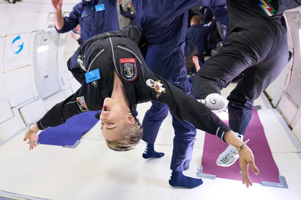 A petite person with short blond hair and light skin stretches out upside down, floating in the zero-g plane, with other bodies floating behind her. Her eyes are closed and mouth open with a slight smile