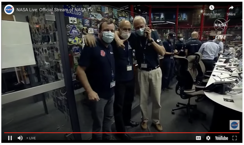 Three men standing with arms around each other, smiling under masks, in mission control