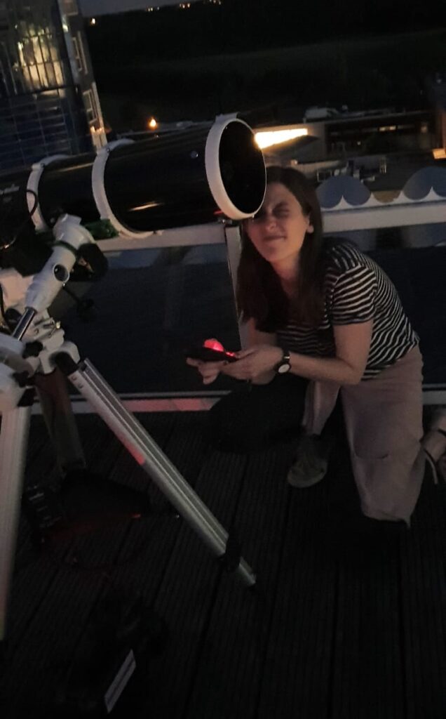 A woman kneels by a telescope, looking into the eye piece. 