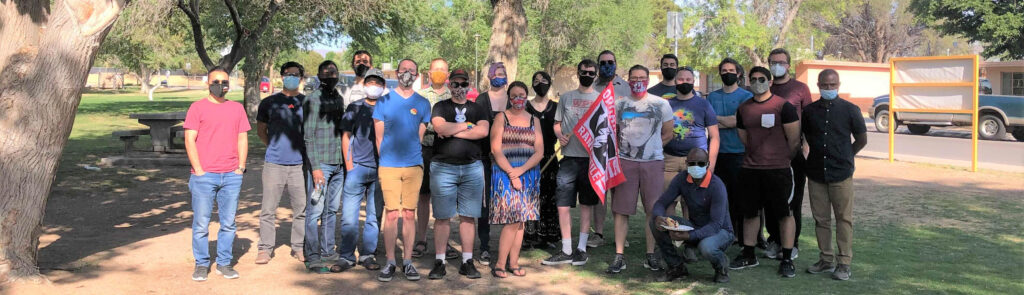 A group of ~20 people in a park with trees. They all have masks on. One is holding a flag that is red and black with text that is not readable. These are members of the organization committee to form a Union at New Mexico State university