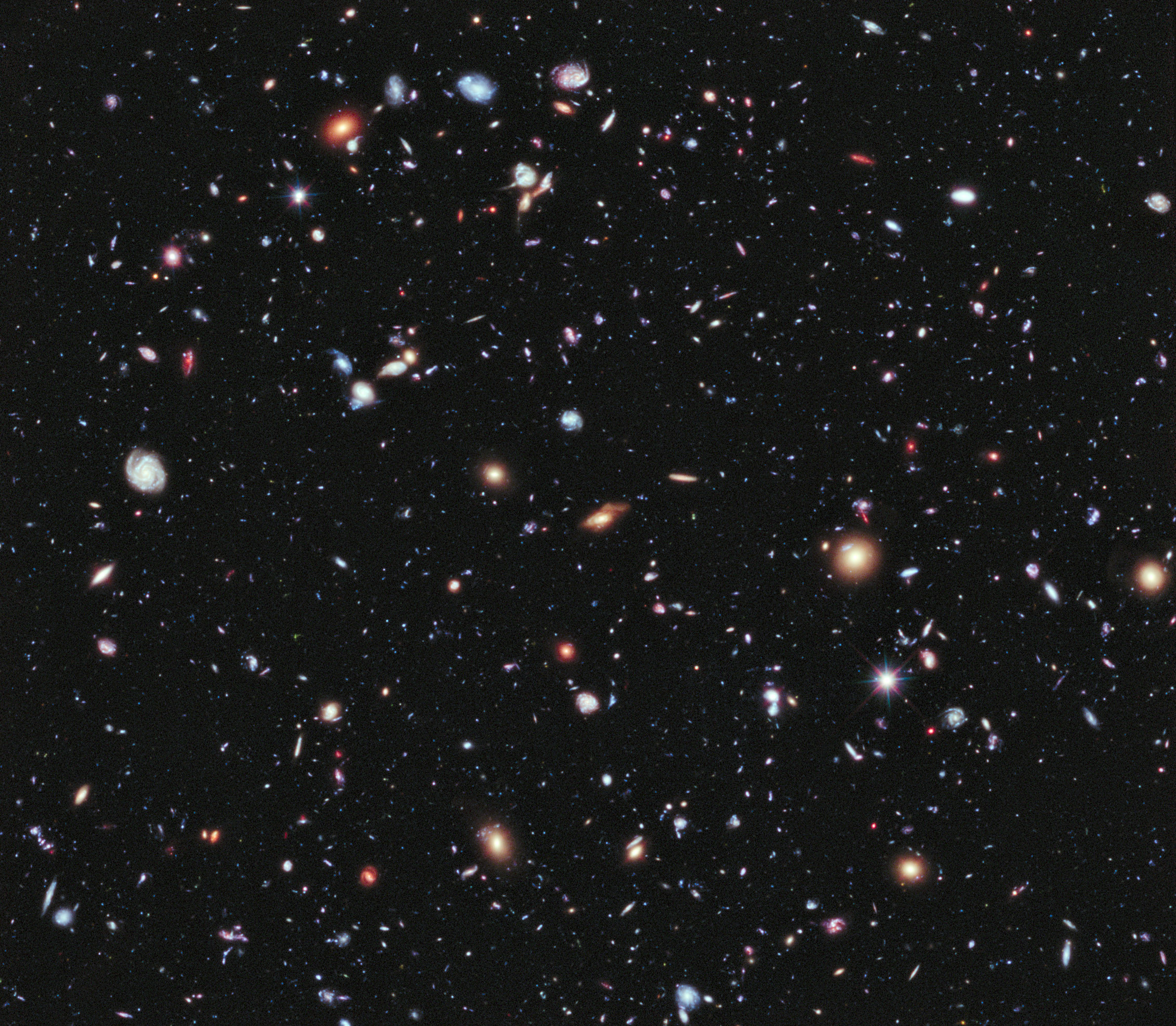 Hubble Space Telescope image showing a field of thousands of galaxies appearing as blobs of different sizes, shapes, and colours, against a black backdrop.