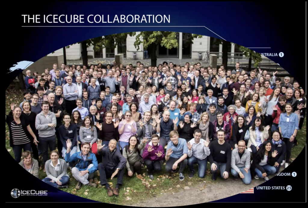 A slide titled "The IceCube Collaboration" with the IceCube logo in the bottom, and an elliptical image of 100s of people sitting or standing on grass in front of a building. They are smiling at the camera which is set high up, and are laughing and waving. 