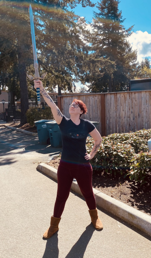 White non-binary person with short red hair in a power pose holding a long sword into the air.