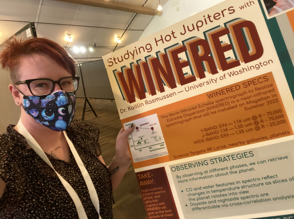 Non-binary person with short red hair and a space-print mask standing in front of a poster titled "Studying Hot Jupiters with WINERED"