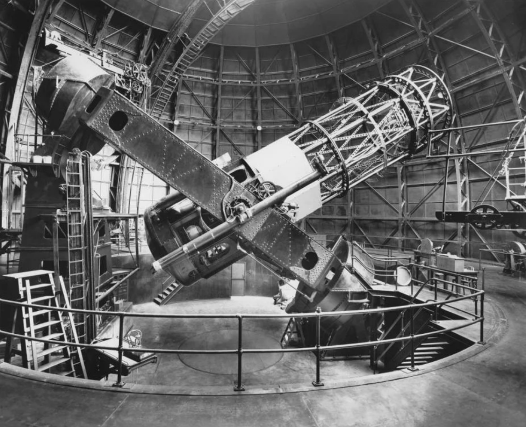 A black-and-white image of the 100-meter Hooker telescope from Mount Wilson, inside its dome. The main part of the telescope looks like a cylinder, mounted on a rotating rectangular platform that gives it two degrees of freedom in which to move.