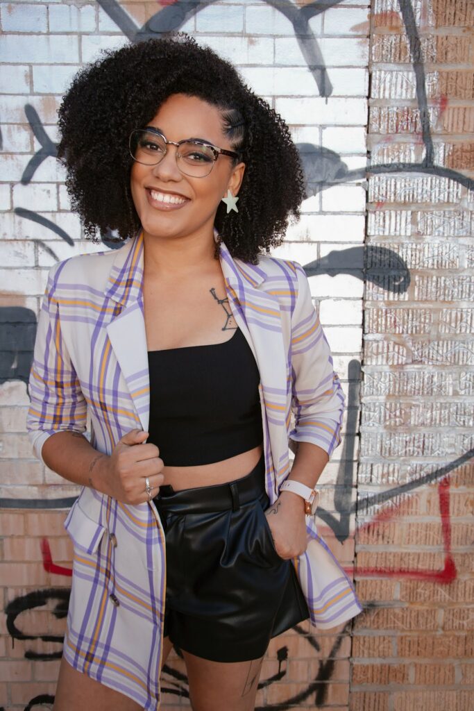 Moiya McTier's author headshot. Image of a Black woman smiling with glasses and large silver star stud earrings, wearing a purple plaid blazer over a black top and black leather shorts, in front of a brick wall with graffiti.
