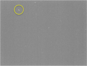 A grey background is dotted with point sources of white light. In the top left, a small trail is seen. The trail is enclosed in a yellow circle to highlight its position
