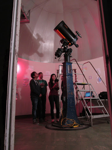 Three people standing in a red-lit room with a telescope in the middle