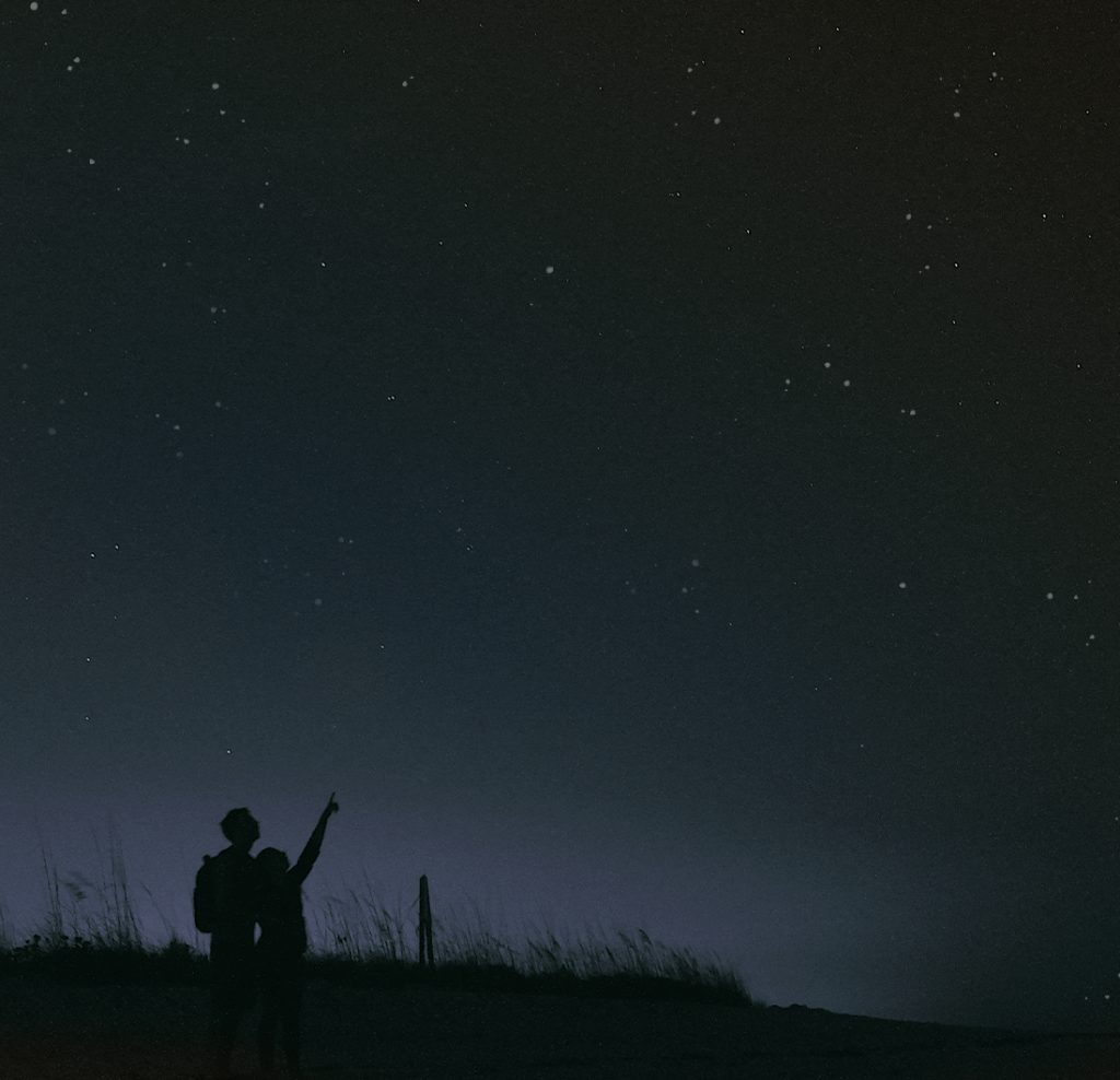 Image is of a couple stargazing. At the lower left corner of the photo there is a small elevated patch of tall grass sitting atop a hill of what could be sand, and the silhouettes of the couple are in front of this hill. As the image moves from left to right the hill flattens and becomes what could be sand. In the lower left side of the image behind the small hill there is a soft light in the background that is a blueish-gray, and the rest of the photo is dark with bright stars dotting the image. The couple is standing very close to each other and their bodies appear to be facing the sky with their backs turned towards the viewer. The person on the right stands shoulder-height to the person on the left, and is pointing up towards the sky. The person on the left is looking up and is wearing a backpack.