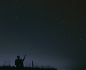 Image is of a couple stargazing. At the lower left corner of the photo there is a small elevated patch of tall grass sitting atop a hill of what could be sand, and the silhouettes of the couple are in front of this hill. As the image moves from left to right the hill flattens and becomes what could be sand. In the lower left side of the image behind the small hill there is a soft light in the background that is a blueish-gray, and the rest of the photo is dark with bright stars dotting the image. The couple is standing very close to each other and their bodies appear to be facing the sky with their backs turned towards the viewer. The person on the right stands shoulder-height to the person on the left, and is pointing up towards the sky. The person on the left is looking up and is wearing a backpack.