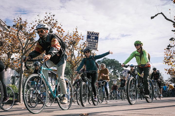 Photo of a large group of strikers riding bicycles on the UC Berkeley campus.  Three runners are highlighted - one on the left looking focused, one in the middle waving at the camera and holding a strike sign, and one on the right - all wearing helmets.