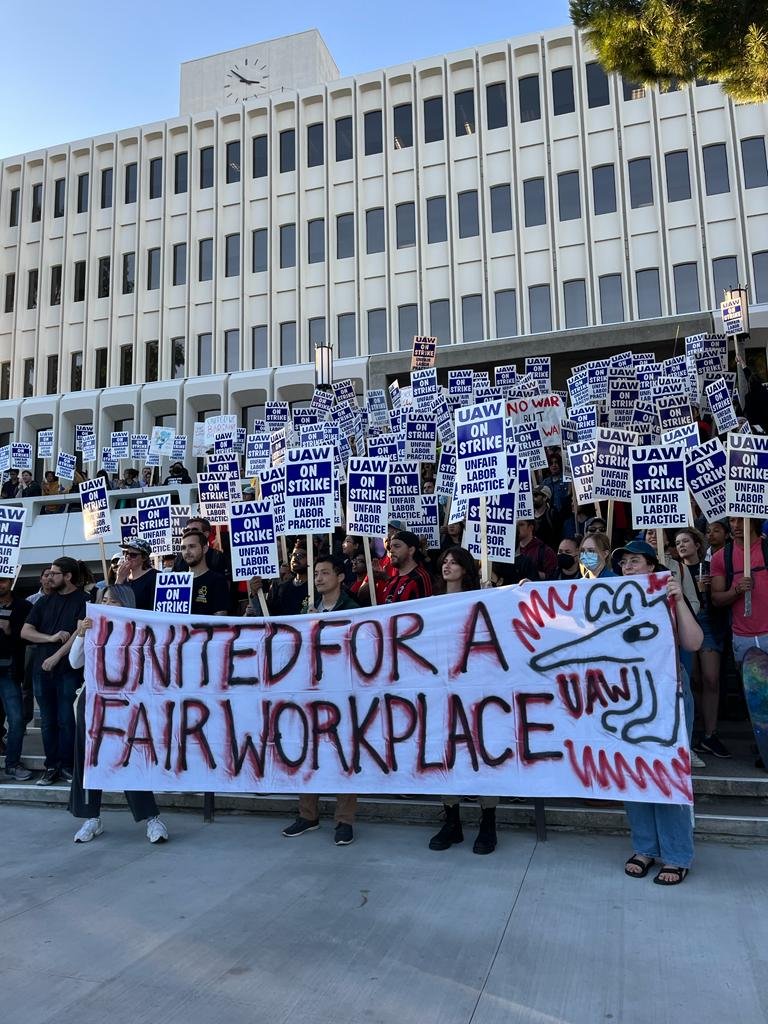 Large crowd of strikers standing outside an academic building at UC Irvine and shouting slogans. Most strikers are holding up placards saying "UAW on strike". Front row folks are holding up a large banner saying "United for a fair workplace".
