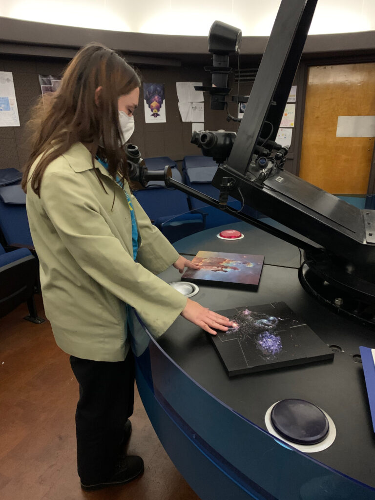 Isabel, a woman with light skin and long dark brown hair, wearing a mask and a green jacket and black pants, touching the two tactile panels as they sit on a platform below the planetarium projector.