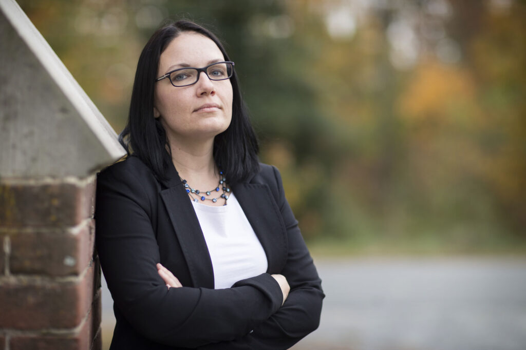 An image of Sabrina Stierwalt. She has black hear and dark eyes, and is wearing a black blazer over a white shirt, a necklace, and black glasses. She is standing with her arms crossed, shoulder resting on the side of a brick structure.