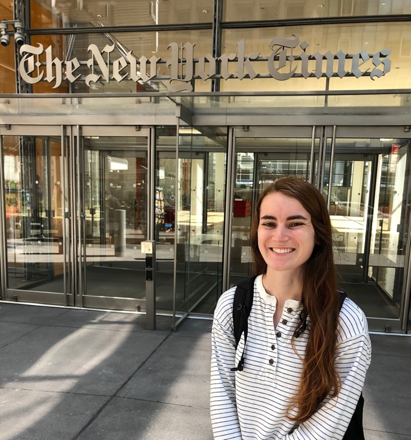 The author of this post standing in front of the New York Times offices. She has long brown hair and is smiling at the camera