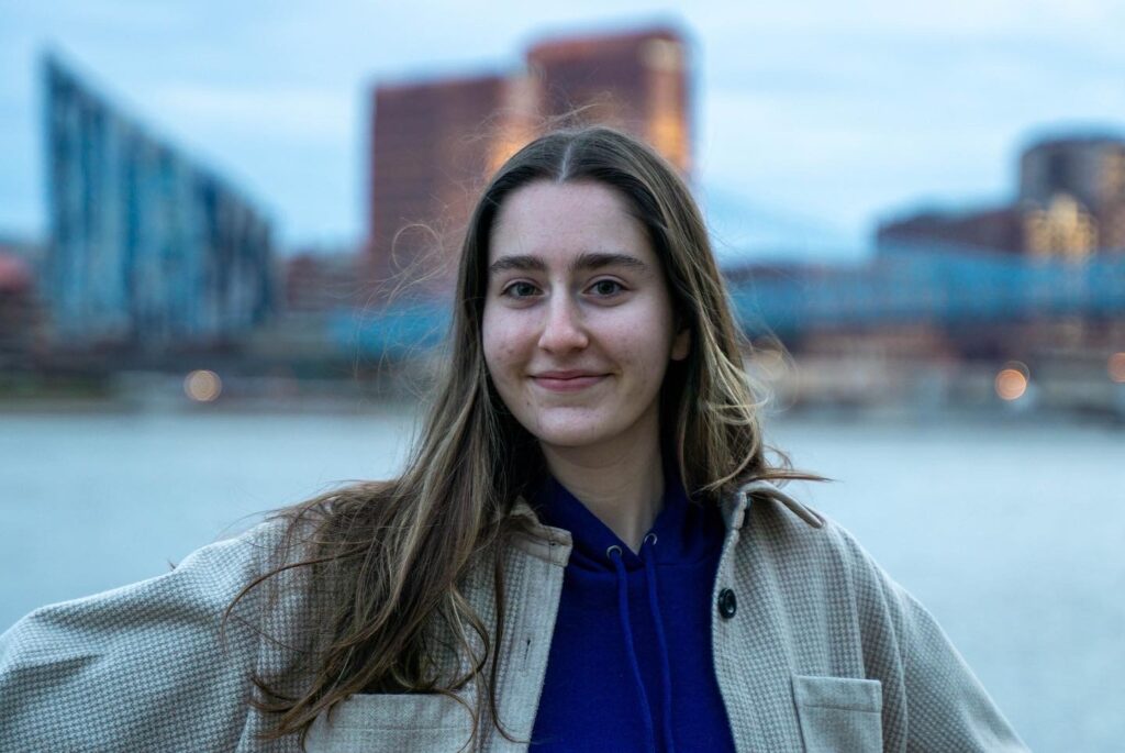 Photo of Lauren Elicker wearing a navy blue hoodie and a beige jacket. A river and tall buildings appear in the background.