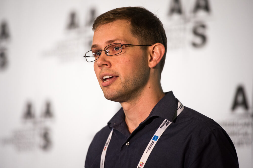 A photo of Brett McGuire taken from a 45 degree angle. He is a caucasian man with short, mildly dark brown har and is wearing glasses and a black collared button-up. He wears a white lanyard around his neck. In the background, the AAS logo is seen in a repeated pattern, blurred.