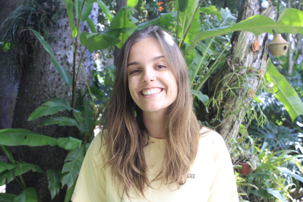 Juliana, a person with long brown hair, smiles widely at the camera. There are broad leaves on plants behind her