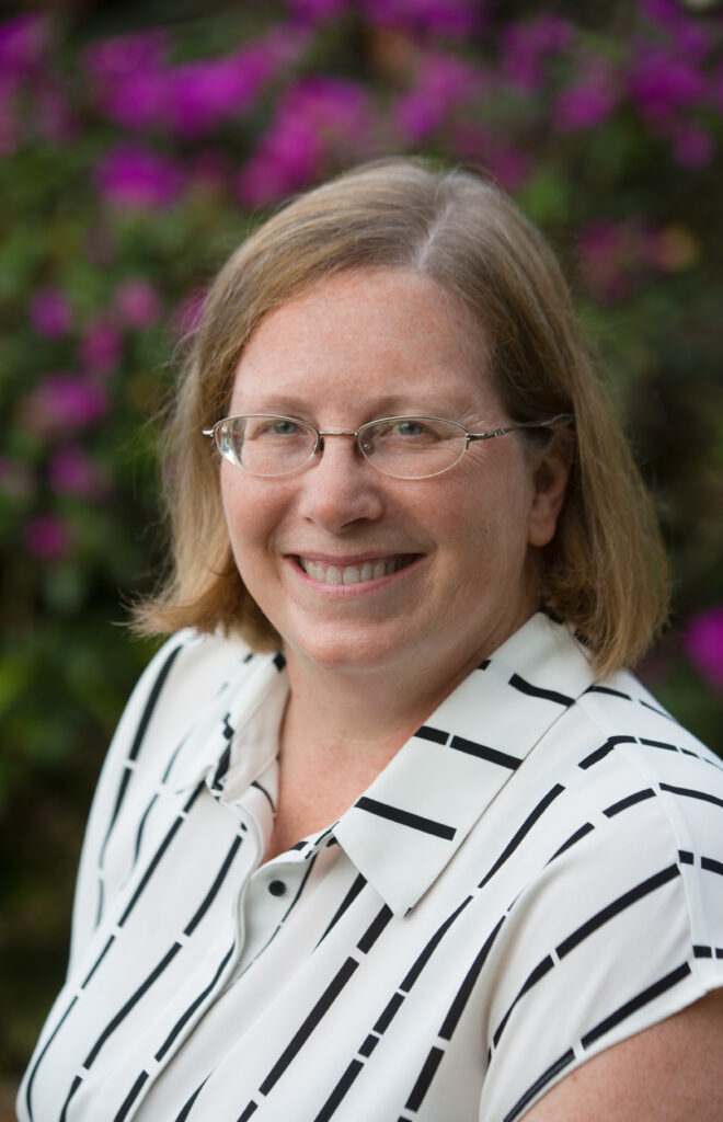 Dr. Karen Meech is pictured. She is a middle-aged caucasian woman with shoulder-length dirty blond hair who wears oval-shaped glasses. She is wearing a black and white stripped collared shirt. In the blurred background, there are magenta flowers.