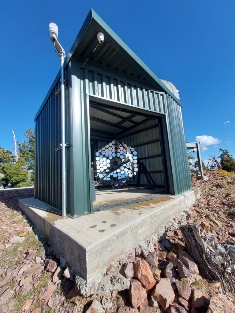 Image of the Trinity Demonstrator. The telescope consists of a hexagonal arrangement of mirrors that direct light to the detectors. It is installed in a shed with a garage-style door that opens and closes depending on whether the Demonstrator is in use.