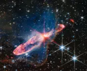At the center is a thin horizontal orange cloud known as Herbig-Haro 46/47 that is uneven with rounded ends, and tilted from bottom left to top right. It takes up about two-thirds of the length of this angle, but is thin at the opposite angle. At its center is a red-and-pink star with prominent, eight-pointed diffraction spikes. It has a central yellow-white blob. The orange lobe to the left is fatter. Just off the edge is a tiny red arc that curves in the opposite direction. The right lobe is thinner, and ends in a smaller orange semi-circle that has a faint purple outline. Just off the edge of this lobe is a slightly smaller orange sponge-like blob. A thin, undulating blue line runs from the central stars through the right lobe. A delicate, semi-transparent blue cloud known as a nebula drifts toward the top of the image and peters out toward the left of the frame, but toward the right and bottom, it ends in a soft ridge set off in a translucent orange. The background is filled with stars and galaxies.