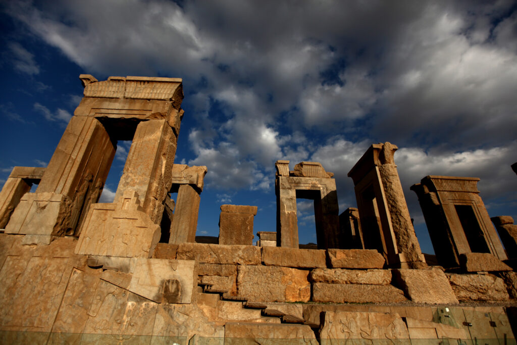 A view of Persepolis, an ancient ruin built of large reddish stones. All that remains are a series of arches, but these are intricately carved. 