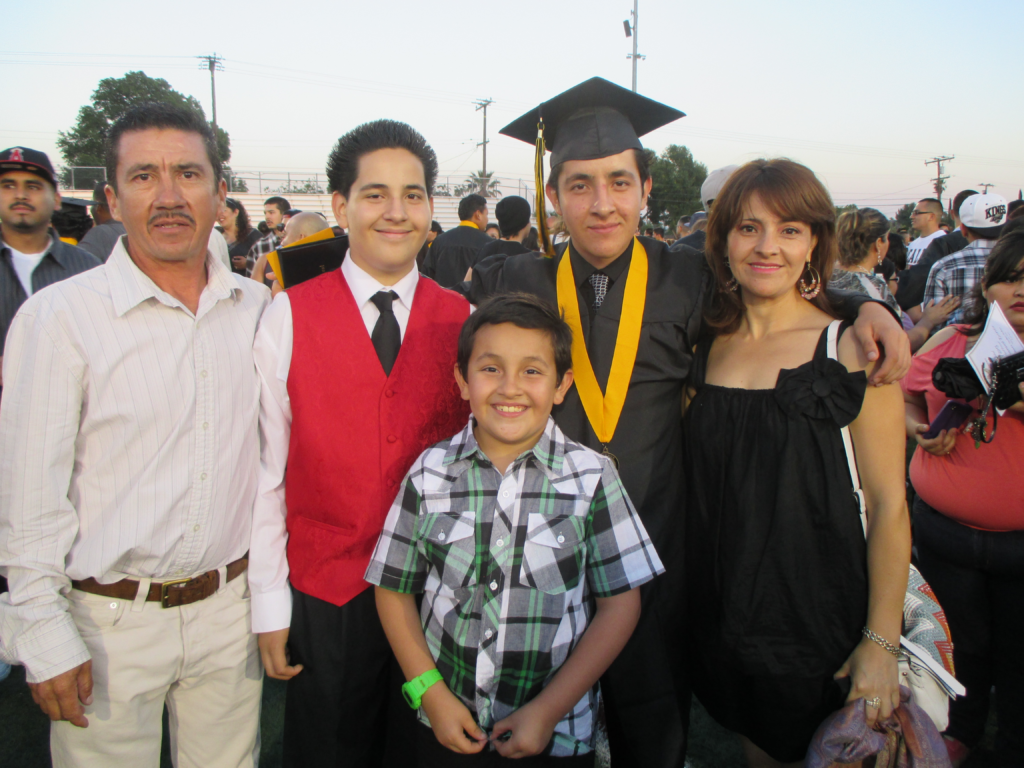 Oscar stands in his graduation gown and cap surrounded by his dad, two younger brothers (off to his left) and his mom (off to his right). Everyone is smiling, especially his youngest brother in the front right!