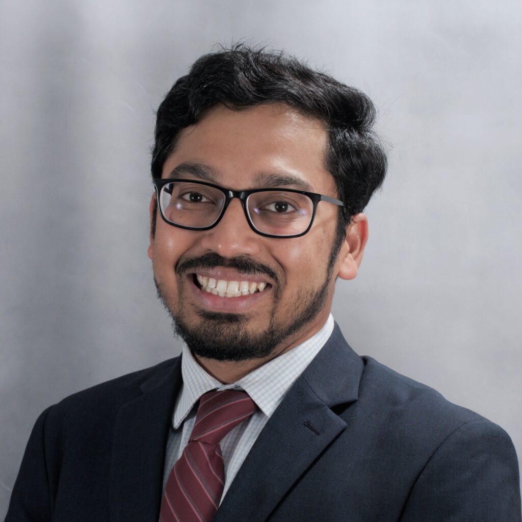 A headshot of Teja, a man with brown skin, black hair, and glasses, who is wearing a suit and tie and smiling for the camera.