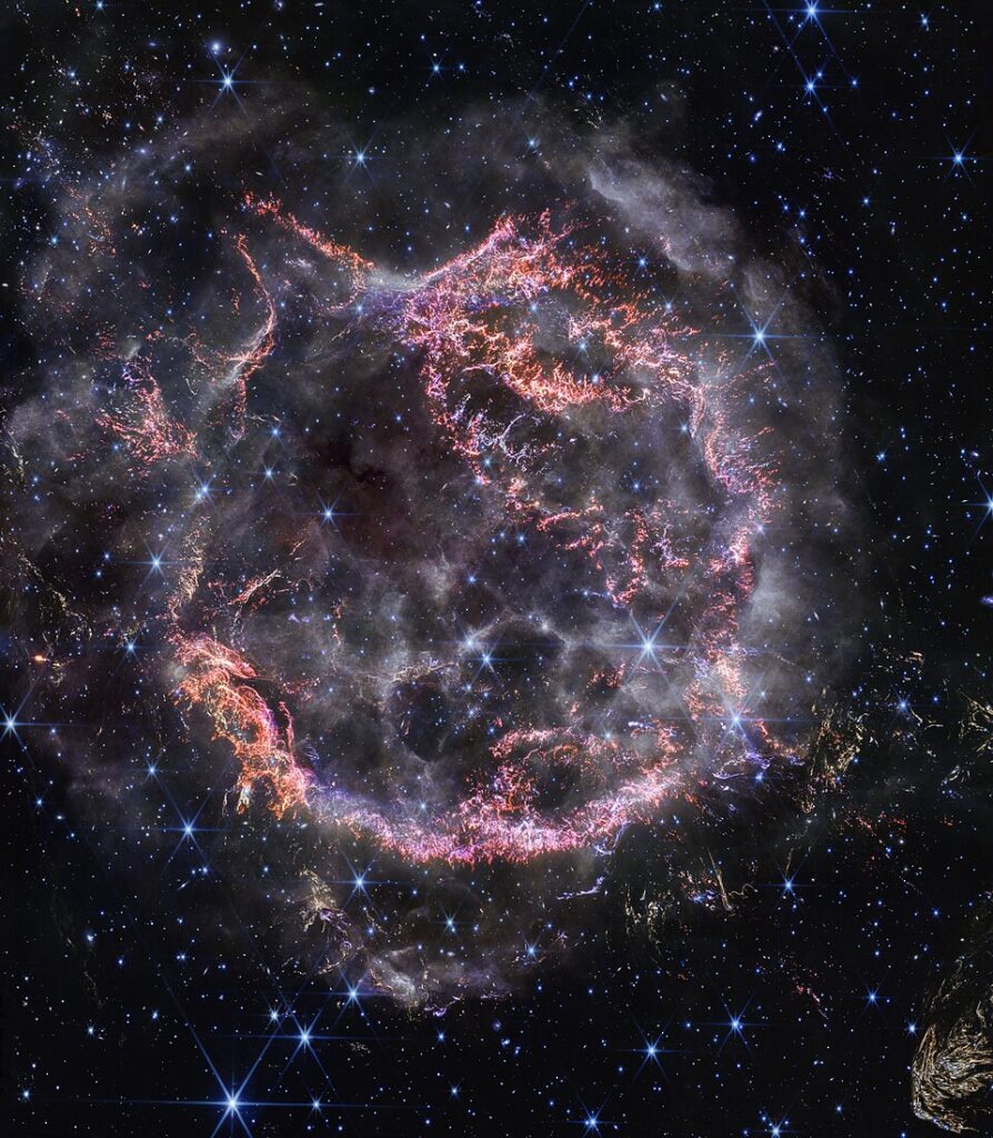 A new high-definition image from the NASA/ESA/CSA James Webb Space Telescope’s NIRCam (Near-Infrared Camera) unveils intricate details of supernova remnant Cassiopeia A (Cas A), and shows the expanding shell of material slamming into the gas shed by the star before it exploded. The most noticeable colours in Webb’s newest image are clumps of bright orange and light pink that make up the inner shell of the supernova remnant. These tiny knots of gas, composed of sulphur, oxygen, argon, and neon from the star itself, are only detectable thanks to NIRCam’s exquisite resolution, and give researchers a hint at how the dying star shattered like glass when it exploded. The outskirts of the main inner shell look like smoke from a campfire. This marks where ejected material from the exploded star is ramming into surrounding circumstellar material. Researchers have concluded that this white colour is light from synchrotron radiation, which is generated by charged particles travelling at extremely high speeds and spiralling around magnetic field lines. There are also several light echoes visible in this image, most notably in the bottom right corner. This is where light from the star’s long-ago explosion has reached, and is warming, distant dust, which glows as it cools down. [Image description: A roughly circular cloud of gas and dust with complex structure. The inner shell is made of bright pink and orange filaments studded with clumps and knots that look like tiny pieces of shattered glass. Around the exterior of the inner shell, there are curtains of wispy gas that look like campfire smoke. Around and within the nebula, various stars are seen as points of blue and white light. Outside the nebula, there are also clumps of dust, coloured yellow in the image.]