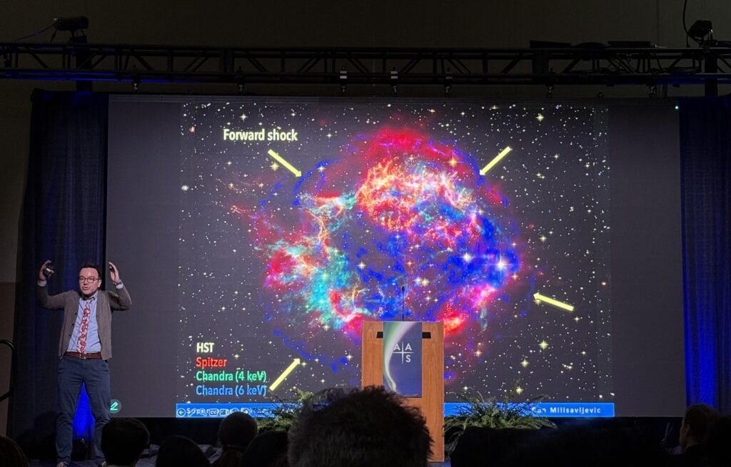 Dan Milisavljevic stands on the left side of the stage while explaining research into the supernova remnant Cassiopeia A, which is displayed on the central screen. The remnant is circular and features many distinct colors tracing out features like a forward shock front.