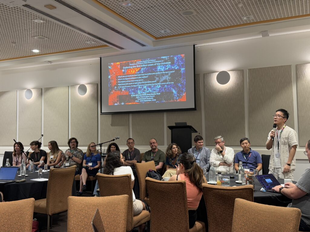 A panel discussion at a conference. About a dozen speakers are seated in a row at the front of a conference room with microphones on stands in front of them. Yuxiang Qin (the main conference organizer) stands on the right holding a microphone and speaking to the audience. Behind them, a projected slide titled “Exploring the first billion years of the Universe” is displayed showing these five questions: 1) What's the one result you most want from first-billion-year studies in the next 5 years?, 2) What's the biggest bottleneck in your subfield?, 3) Reionization will be solved when we xxx, 4) What keeps you awake at night about the first billion years?, 5) In the next 10 years, the biggest advance in studying the first billion years will be xxx. Audience members sit facing the panel, some with laptops open in a well-lit room with beige walls and brown chairs.
