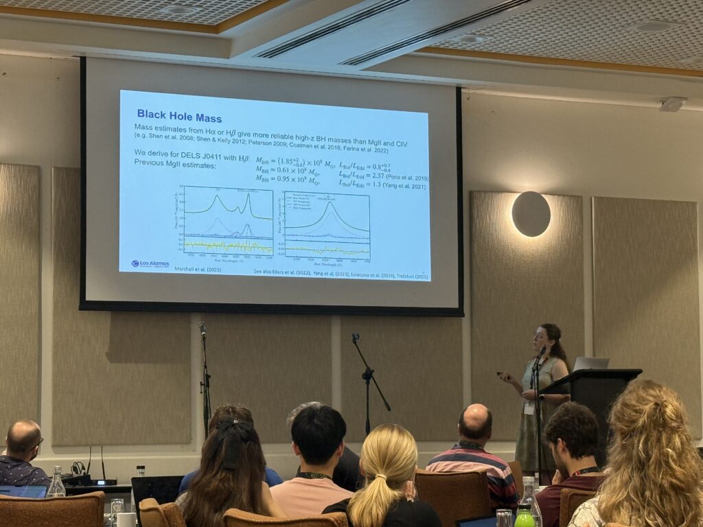 Madeline Marshall stands at a podium delivering a talk with a slide titled “Black Hole Mass”. The slide compares spectral fits of quasar emission lines (Hβ/Halpha and Mg II/CIV) and lists derived black hole masses and Eddington ratios for the quasar DELS J0411. Plots show emission line profiles and model fits, illustrating differences between previous and updated mass estimates. 
