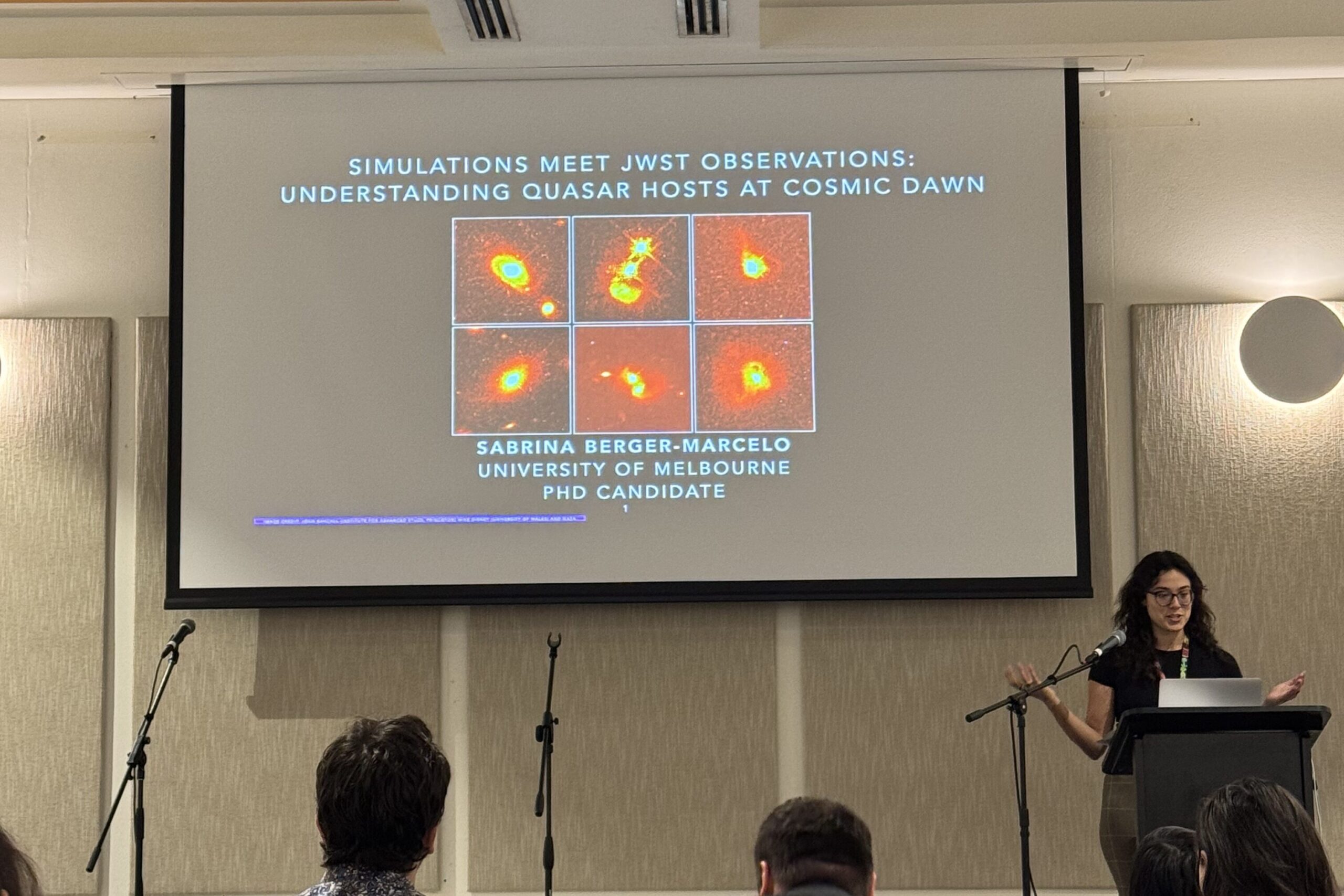 Sabrina Berger standing at a podium in front of an auditorium. Behind her a slide is projected onto a screen that is mostly cut off by the photograph. The slide contains three side by side images of white and yellow quasars and their host galaxies on an orange background. Beneath is text that reads “Sabrina Berger-Marcelo, University of Melbourne, PhD Candidate”.

