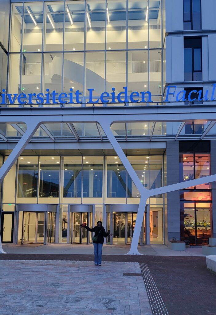 Flore Milien standing in front of the University of Leiden building. The building has a lot of glass windows and gray metal structure with the name of the university in blue letters. She's standing far in the background wearing a balck jacket, green shoulder bag and blue jeans