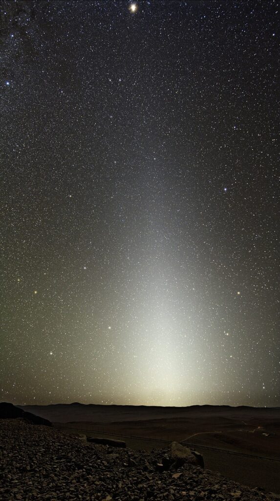 Zodiacal light as seen from Cerra Paranal. The light is a bright white triangle that slowly fades into the background of the dark sky filled with stars, with dark mountains on the horizon